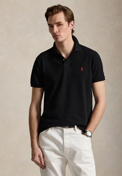 Junger Mann trägt ein schwarzes Polohemd mit rotem Logo und weißen Hosen, steht vor einem schlichten beigen Hintergrund.