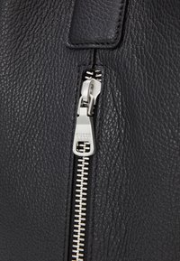 Borsa a mano in pelle nera con finitura testurizzata, dotata di una cerniera argentata con branding. La cucitura dettagliata completa il design della cerniera.