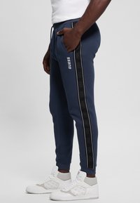 Pantaloni felpati blu navy con strisce laterali nere, caratterizzati da un motivo a logo, cintura elasticizzata e polsini. Abbinati a sneaker alte bianche.