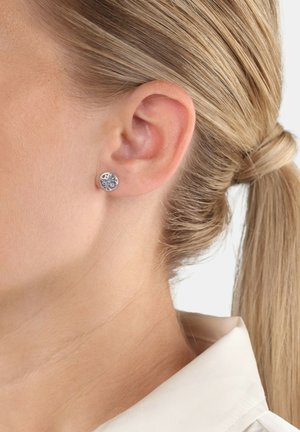 Pendiente de plata con un diseño de árbol, llevado en la oreja izquierda de una persona con el cabello rubio recogido en una coleta.
