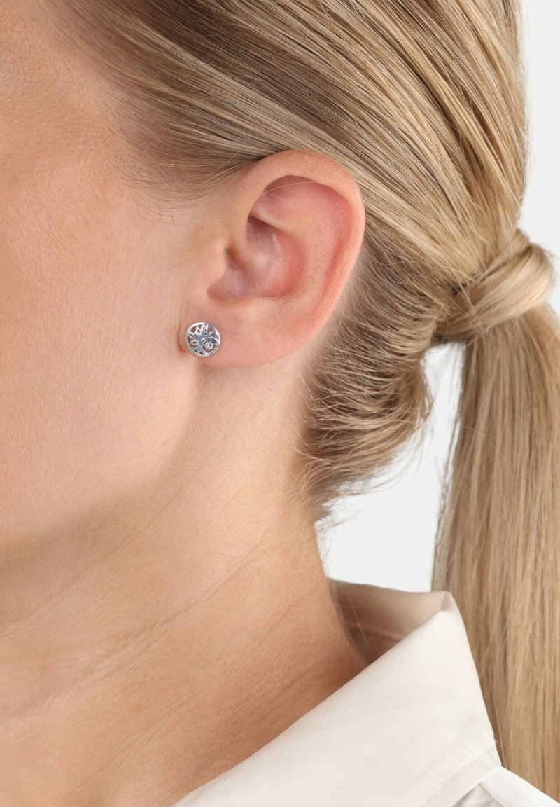 Pendiente de plata con un diseño de árbol, llevado en la oreja izquierda de una persona con el cabello rubio recogido en una coleta.