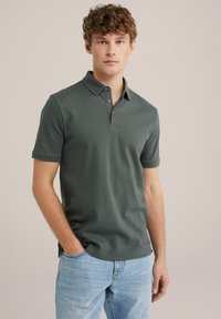 Polo shirt de color verde oscuro hecho de algodón suave, con una tapeta de tres botones y mangas cortas. Combinado con unos jeans de color azul claro.