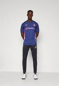 Blue athletic shirt with pink accents, short sleeves, and logo; paired with black pants and white sneakers. Smooth fabric, modern fit.