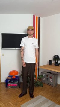 Bílá tričko s potiskem vpředu, hnědé kalhoty a béžová čepice. Stojí v místnosti s televizí, barevnými pruhy na stěně a dekoračními předměty.