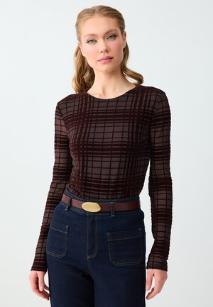 Vrouw met lichtbruin haar die een doorschijnende top met lange mouwen en een donker ruitpatroon draagt, donkerblauwe broek en een bruine riem met een gouden gesp.