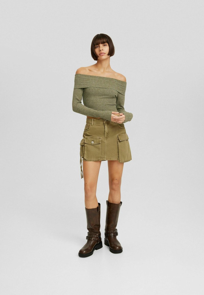 Top off-shoulder a coste verde oliva abbinato a una mini gonna khaki con tasche laterali, completato da stivali marroni fino al ginocchio.