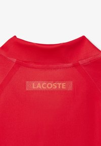 Röd polo-shirt med rundad krage och en texturerad, klar logotyp på baksidan. MATERIALET verkar slätt och figursytt.