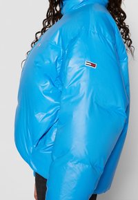 Blue puffer jacket with a glossy finish, high collar, and embroidered logo detail on the sleeve. Features a loose, oversized fit.