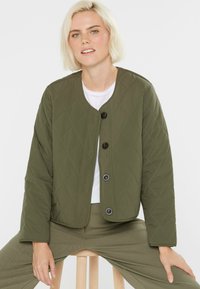 Blonde Frau, die auf einem Holzschemel sitzt und eine olivgrüne gesteppte Jacke mit Knöpfen über einem weißen Shirt und passenden Hosen trägt.