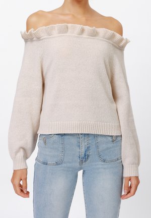 Frau trägt einen beigefarbenen, schulterfreien Pullover mit Rüschensaum und hellblaue, hoch taillierte Jeans mit sichtbaren Vordertaschen.