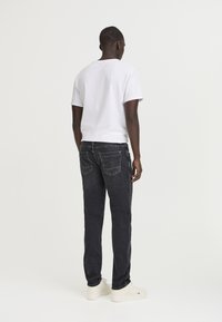 Men's white short-sleeve t-shirt and dark denim jeans. The jeans feature back pockets and subtle stitching. The shoes are white with minimal branding.