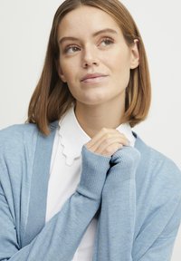 Světle modrý cardigan s žebrovanou texturou a přiléhavými manžetami, nošený přes bílou košili s výstřihem ve tvaru vlny. Minimalistický design, měkký materiál.