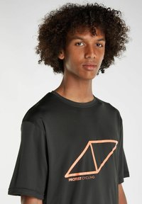 Mustapaita pyöräilyyn, jossa on korallinvärinen geometrinen logo etupuolella, valmistettu hengittävästä materiaalista, jossa on teksturoituvat hihat ja pyöreä kaula-aukko.