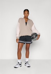 Lichtbeige ritsjack met lichtroze mouwen, gecombineerd met zwarte shorts met witte strepen. Houdt een basketbal vast. Witte sneakers en sokken.
