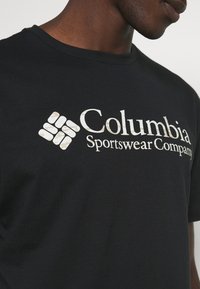 T-shirt noir avec le logo blanc de Columbia Sportswear Company porté par une personne à la peau foncée et à la mâchoire visible.