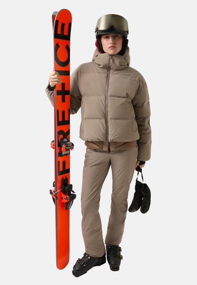 Sprzęt narciarski składający się z brązowej ocieplanej kurtki, brązowych spodni narciarskich, czarnych butów narciarskich i pomarańczowych nart z czarnymi napisami, trzymanych razem z czarnymi rękawiczkami.