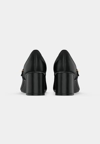 Pár čiernych kožených topánok na blokovom opätku so zlatými sponami zobrazených zozadu na jednoduchom bielom pozadí.