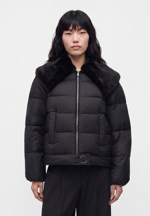 Un mannequin aux longs cheveux noirs porte une doudoune noire matelassée avec un large col en fausse fourrure, une fermeture éclair sur le devant, des poches et une sangle à boucle.