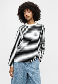 Μακρυμάνικη μπλούζα με μαύρες και λευκές ρίγες, στρογγυλή λαιμόκοψη, απαλό ύφασμα και μικρή λεπτομέρεια λογότυπου στο στήθος. Συνδυασμένη με ανοιχτό μπλε παντελόνι με σχέδιο.