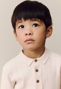 Maglietta polo rosa chiaro lavorata a maglia con un motivo a trecce, caratterizzata da quattro bottoni marroni e un colletto classico, indossata da un bambino con capelli corti e scuri.