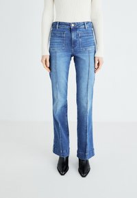 Jeans flare in denim blu con vita alta, cinque tasche e cuciture decorative. Indossati con stivali neri a punta.