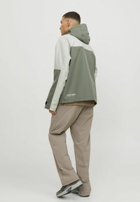 Mandens farveblokerede jakke i olivengrøn og lysegrå, med hætte, iført beige bukser og grå sneakers. Teksturstof, afslappet pasform.