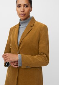 Mosterdkleurige ribfluwelen blazer met pieklapels en een enkele knoopsluiting, gedragen over een lichtgrijze coltrui. Getextureerde stof.