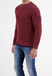 Bordeauxfärgad stickad tröja med ribbade ärmslut och nederkant, med ett texturerat mönster och en liten blå logotyp på vänster ärm.