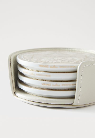 Sada čtyř bílých keramických podtácků s béžovým barokním vzorem, uložených v světle béžovém koženém obalu s prošitými detaily. Obsahuje značku Versace.