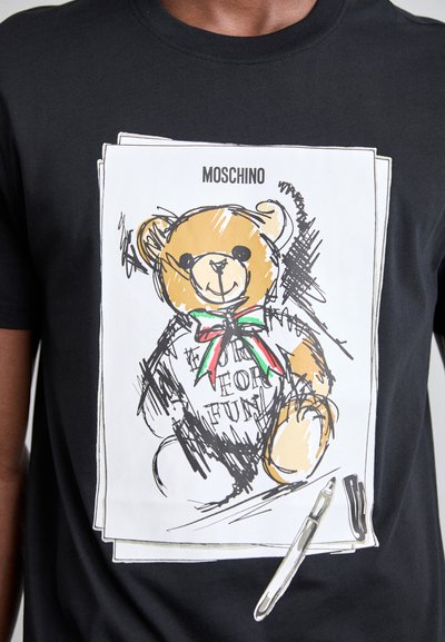 T-shirt noir avec un graphique de ours en dessin animé, contourné en noir, avec un ours marron clair et un ruban coloré. Le texte indique "POUR LE PLAISIR".