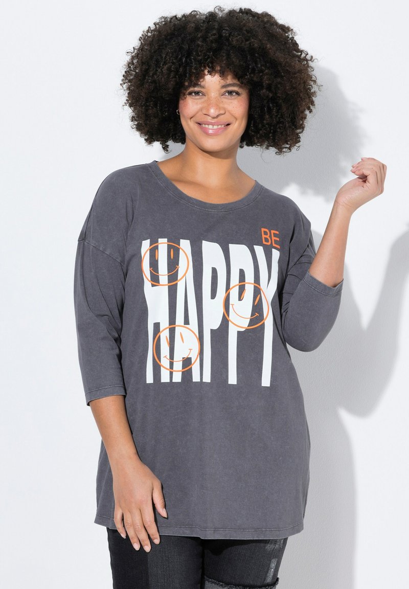Grijze oversized T-shirt met drie oranje lachende gezichten en vet wit tekst die "WEES VROLIJK" zegt. Heeft driekwart mouwen en zachte stof.