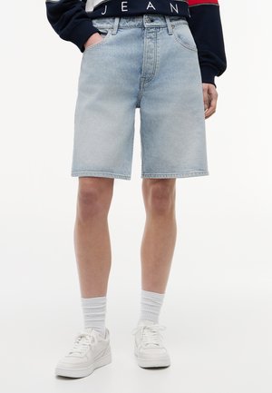 Osoba ubrana w jasnoniebieskie szorty dżinsowe, białe skarpetki, białe trampki i granatową bluzę z napisem "JEAN" na dole.