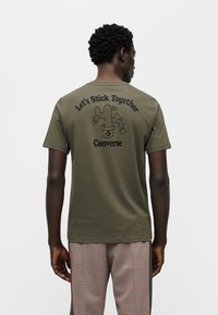 Χακί πράσινη βαμβακερή μπλούζα με γραφικό κάκτου και κείμενο στην πλάτη. Κοντά μανίκια και σχέδιο με στρογγυλή λαιμόκοψη, συνδυασμένη με καρό παντελόνια.