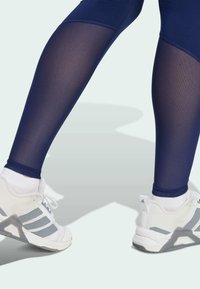 Leggings de sport bleu marine avec des panneaux transparents, associés à des baskets blanches avec des accents et des lacets gris, mettant en valeur un design sportif.