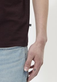 Camiseta marrón de algodón, con una etiqueta lateral y jeans de mezclilla azul en contraste. Primer plano de una mano descansando sobre los jeans.