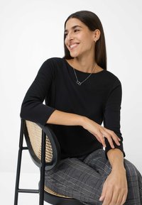 Zwarte lange mouwen top gecombineerd met grijze geruite broek; model zittend op een zwarte stoel met een geweven rugleuning. Zilveren ketting met ronde hangers.