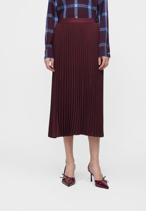Femme portant une jupe midi plissée bordeaux, des chaussures à talons hauts pointues bordeaux avec des nœuds, et une chemise à carreaux bleue et bordeaux boutonnée.
