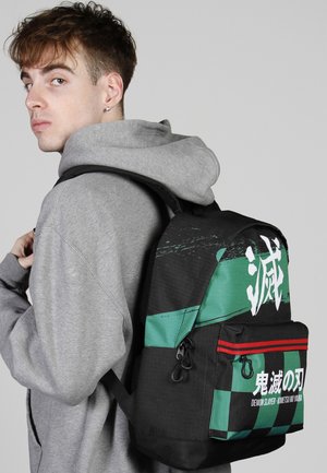 Jonge persoon in een grijze hoodie met een zwarte en groene Demon Slayer-rugzak met Japanse karakters en rode strepen over één schouder.