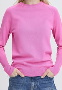 Une personne portant un pull long à manches longues de couleur rose vif avec des poignets côtelés, associé à un jean en denim bleu, les mains glissées dans les poches avant.