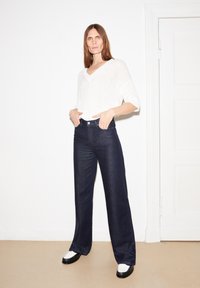 Maglione bianco a maglia con scollo a V, abbinato a jeans a gamba larga blu scuro a vita alta. Scarpe platform bianco e nere. Sfondo semplice.