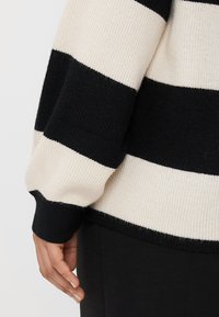 Svart og kremfargede striper på en genser med ribbet tekstur, som har en løs passform og mansjetterte ermer. Bunnkanten er også svart.