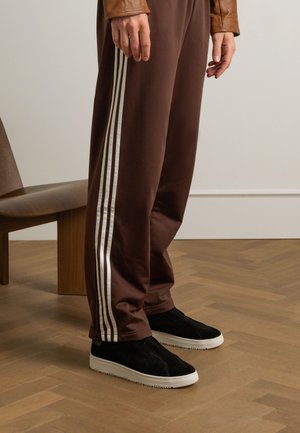 Brown athletic pants with white stripes, paired with black suede slip-on shoes featuring a white rubber sole, against a wooden floor.