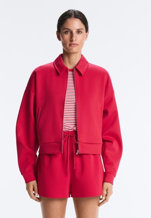 Vrouw die een rood jasje met rits draagt over een rood-wit gestreept shirt, gecombineerd met rode short met trekkoord, voor zich uit kijkend.