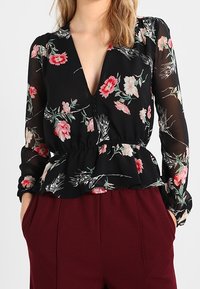 Donna che indossa una blusa nera floreale trasparente con maniche lunghe e bottoni, abbinata a pantaloni bordeaux a vita alta con tasche.