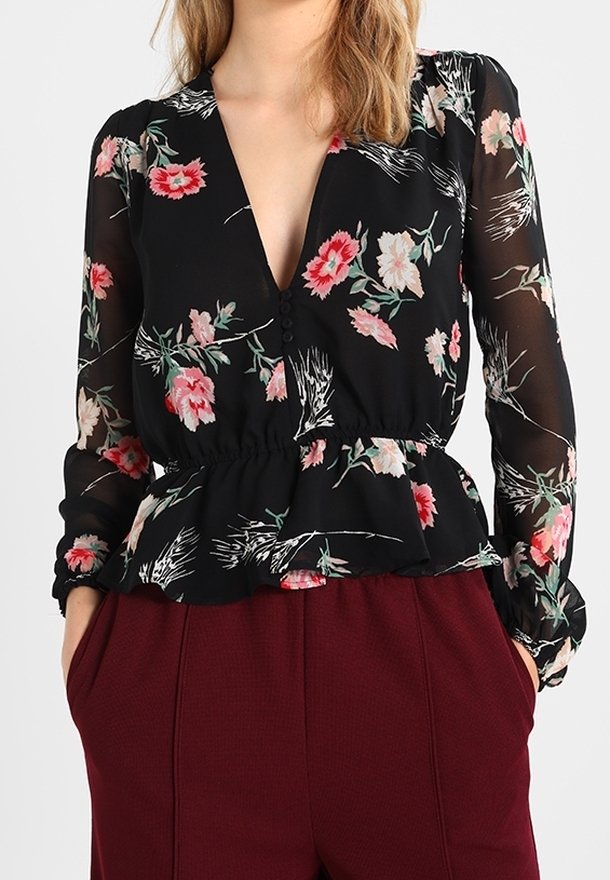 Donna che indossa una blusa nera floreale trasparente con maniche lunghe e bottoni, abbinata a pantaloni bordeaux a vita alta con tasche.