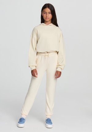 Felpa beige con cappuccio regolabile e vita elastica, abbinata a pantaloni sportivi a turbo coordinati. Il modello indossa scarpe da ginnastica blu. Texture liscia.