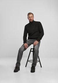 Juodas raštuotas zip-up megztinis, pilkos džinsai, juodi suedinės bateliai, sėdintis ant juodo suolo prieš baltą foną, demonstruojantis kasdienį aprangos stilių.