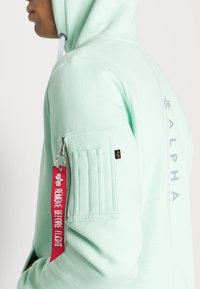 Hoodie verde menta con tasca con zip sulla manica, caratterizzato da un'etichetta rossa "RIMUOVERE PRIMA DEL VOLO" e il logo "ALPHA" sulla parte posteriore.
