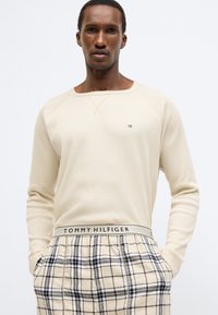 Krém színű kötött pulóver kerek nyakkivágással és hosszú ujjakkal, bézs kockás kényelmi nadrággal, melynek derékpántján márkajelzés található.