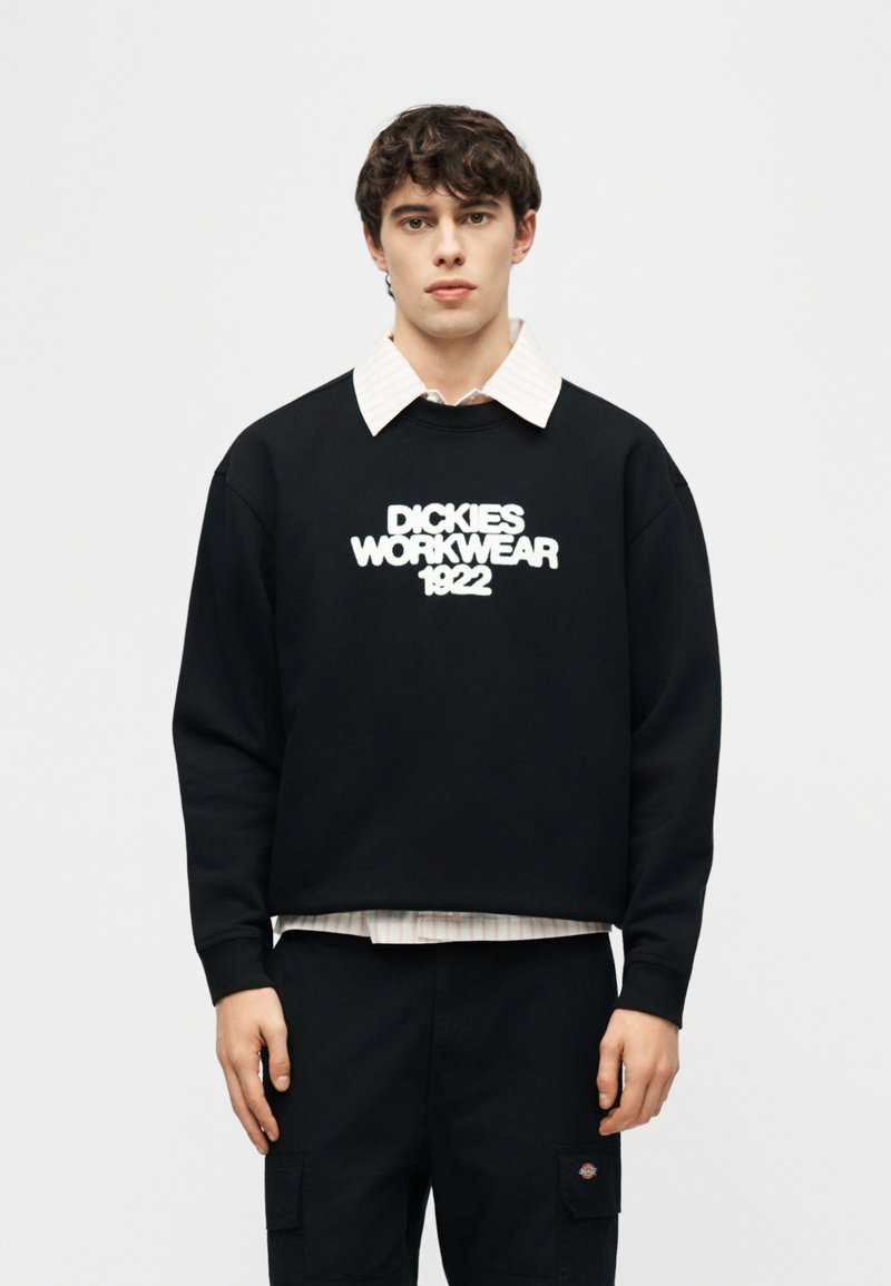 Giovane uomo che indossa una felpa nera con la scritta "DICKIES WORKWEAR 1922", stratificata sopra una camicia bianca con collo e pantaloni neri con tasche cargo.